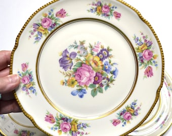 May include: A round porcelain plate with a scalloped edge trimmed in gold. The center features a colorful floral bouquet of pink roses, purple irises, and yellow blossoms. The rim is decorated with smaller matching floral clusters and gold bands.
