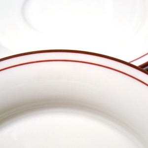 May include: Close-up of white plates with a thin, dark red border. The plates are stacked, showcasing the clean, simple design. The image highlights the smooth, glossy surface and the contrasting colors.