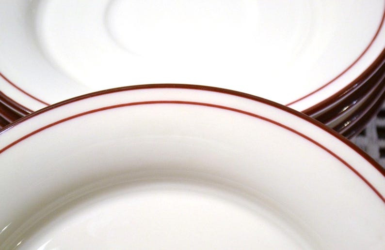 May include: Close-up of white plates with a thin, dark red border. The plates are stacked, showcasing the clean, simple design. The image highlights the smooth, glossy surface and the contrasting colors.