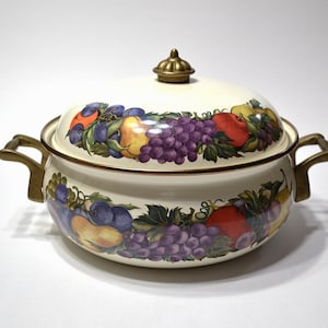 May include: A cream-colored enameled metal casserole dish with a matching lid, featuring a colorful fruit pattern of grapes, pears, and apples. The pot has two brass-toned metal side handles and a matching brass-toned knob on the lid.
