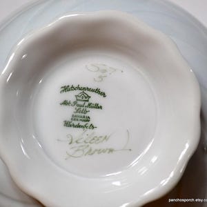 May include: Close-up of a white porcelain bowl's interior, showcasing the maker's mark and signature. The bowl has a scalloped edge and a light blue, swirled design on the exterior. The text includes "Hutschenreuther" and "Eileen Brown."