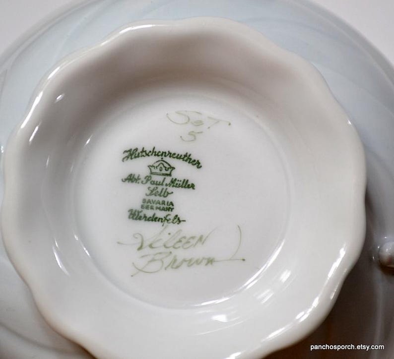 May include: Close-up of a white porcelain bowl's interior, showcasing the maker's mark and signature. The bowl has a scalloped edge and a light blue, swirled design on the exterior. The text includes "Hutschenreuther" and "Eileen Brown."