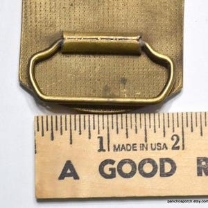 May include: A vintage brass belt buckle with a rectangular shape and a textured surface. The buckle has a curved handle and is next to a wooden ruler that reads "MADE IN USA".