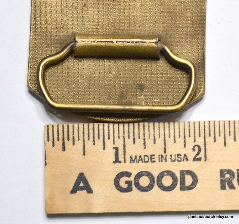May include: A vintage brass belt buckle with a rectangular shape and a textured surface. The buckle has a curved handle and is next to a wooden ruler that reads "MADE IN USA".