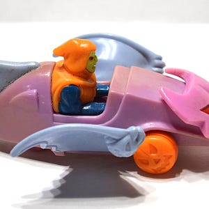 May include: A vintage toy car in shades of pink, blue, and orange, featuring a character figure. The car has a pumpkin-shaped wheel and a jack-o'-lantern design on the front. The character is wearing an orange hood and blue clothing.