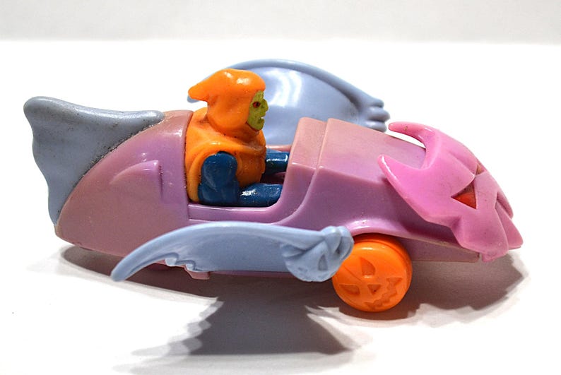 May include: A vintage toy car in shades of pink, blue, and orange, featuring a character figure. The car has a pumpkin-shaped wheel and a jack-o'-lantern design on the front. The character is wearing an orange hood and blue clothing.