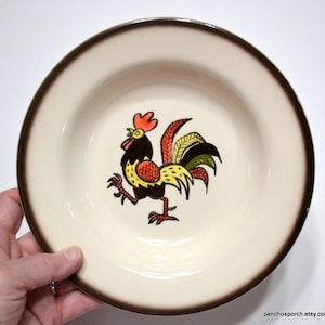 May include: A white ceramic soup bowl with a brown rim features a colorful rooster illustration. The rooster is yellow, red, green, and black.