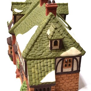 May include: A miniature ceramic house with a green tiled roof, brick walls, and snow accents. The house features multiple windows, chimneys, and a small evergreen tree. The color palette includes green, brown, white, and red.