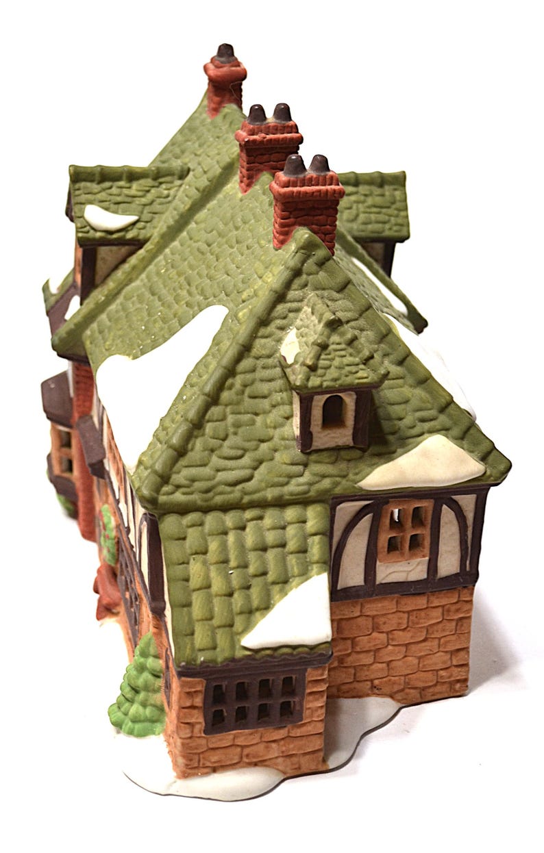 May include: A miniature ceramic house with a green tiled roof, brick walls, and snow accents. The house features multiple windows, chimneys, and a small evergreen tree. The color palette includes green, brown, white, and red.