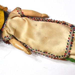 Puede incluir: Mu&ntilde;eca vintage con una cara amarilla en forma de m&aacute;scara, cabello negro y un vestido color crema. El vestido est&aacute; adornado con cuentas de colores y tela verde. Los brazos y las piernas de la mu&ntilde;eca son de madera.