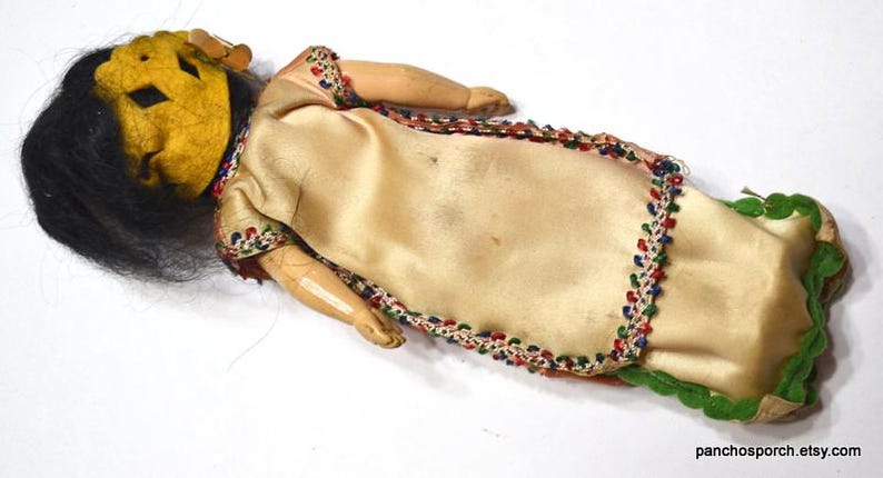 Puede incluir: Mu&ntilde;eca vintage con una cara amarilla en forma de m&aacute;scara, cabello negro y un vestido color crema. El vestido est&aacute; adornado con cuentas de colores y tela verde. Los brazos y las piernas de la mu&ntilde;eca son de madera.