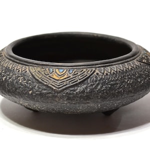 May include: A dark gray, low, round bowl with a textured exterior and three small feet. The rim features a decorative band with geometric patterns in gold and blue. The bowl is likely made of ceramic or stone.