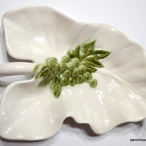 May include: A white ceramic leaf-shaped serving dish with a raised green grape cluster and leaves design. The dish has a handle and is suitable for serving appetizers or snacks.