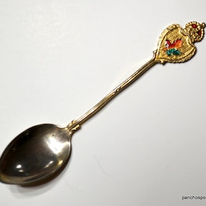 May include: A vintage souvenir spoon with a gold-colored handle and a silver-colored bowl. The spoon's handle features a gold-colored crest with a red, orange, and green maple leaf design, topped with a crown. The spoon is likely a collectible item.
