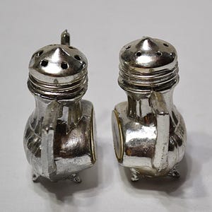 May include: A pair of silver-colored salt and pepper shakers. Each shaker has a handle, a small spout, and a decorative design. The shakers are on a white background.