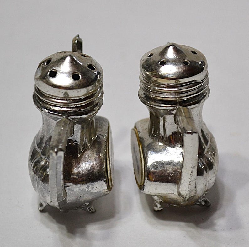 May include: A pair of silver-colored salt and pepper shakers. Each shaker has a handle, a small spout, and a decorative design. The shakers are on a white background.