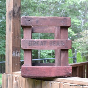 May include: A vintage wooden crate, painted a deep red, with the words "GREAT BEAR" stencilled on the front. The crate is constructed from wooden slats and appears weathered. It is resting on a wooden surface, with a natural background.