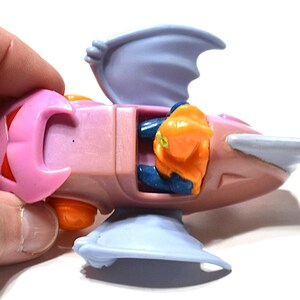 May include: A pink and light blue toy spaceship with orange and blue accents. The toy has a wing-like structure on the top and bottom, and a small orange figure is visible inside the cockpit.