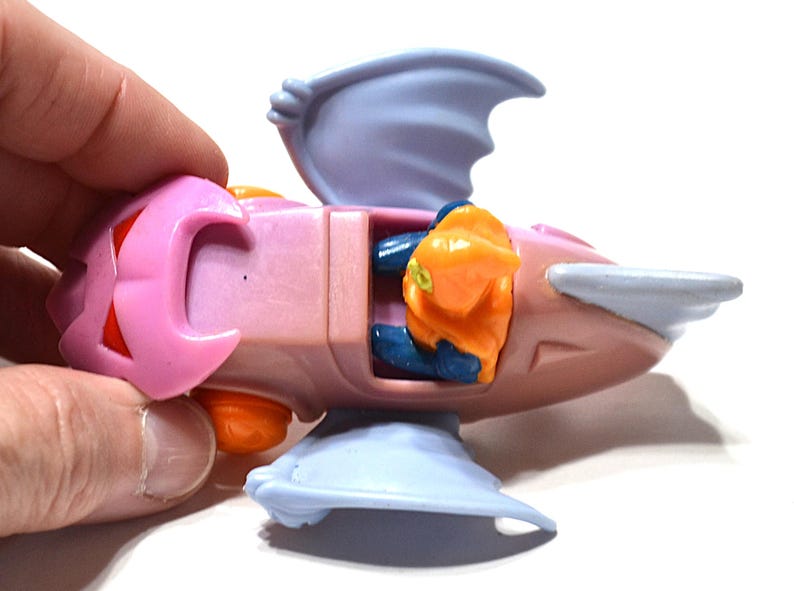 May include: A pink and light blue toy spaceship with orange and blue accents. The toy has a wing-like structure on the top and bottom, and a small orange figure is visible inside the cockpit.