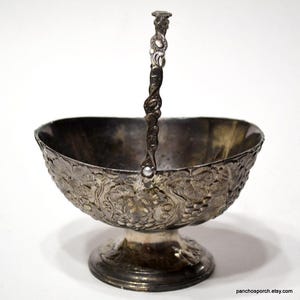 May include: A silver sugar bowl with a handle and an ornate floral design. The bowl is on a pedestal base.