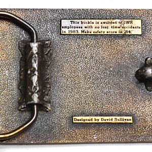 May include: A rectangular metal belt buckle with a textured surface and a gold-coloured frame. The buckle has text that reads "This buckle is awarded to JWR employees with no lost time accidents in 1983. Make safety score in '84." and "Designed by David Sullivan."