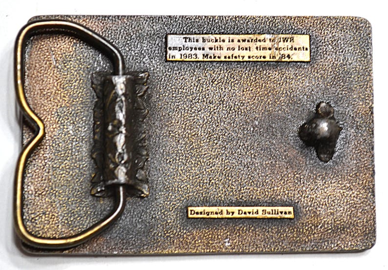 May include: A rectangular metal belt buckle with a textured surface and a gold-coloured frame. The buckle has text that reads "This buckle is awarded to JWR employees with no lost time accidents in 1983. Make safety score in '84." and "Designed by David Sullivan."
