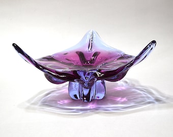 May include: A decorative glass bowl featuring a vibrant purple center that transitions into a lighter lavender hue at the edges. The bowl has a distinct, organic, multi-pointed shape with a thick, molded base that supports the flared, wavy rim.
