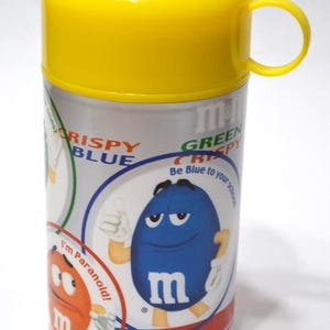 May include: A cylindrical container with a yellow lid and handle, featuring a silver body with colorful graphics. The graphics include the text "CRISPY BLUE" and "GREEN CRISPY", along with cartoon characters and the letter "m".