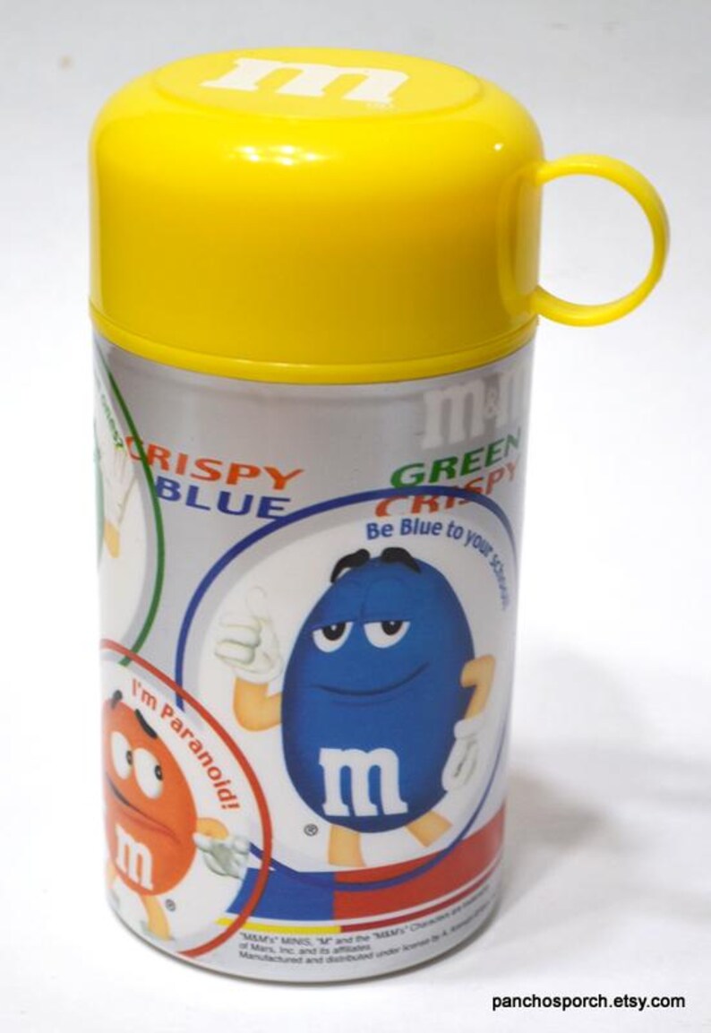 May include: A cylindrical container with a yellow lid and handle, featuring a silver body with colorful graphics. The graphics include the text "CRISPY BLUE" and "GREEN CRISPY", along with cartoon characters and the letter "m".