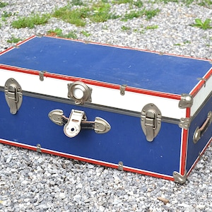 May include: A rectangular storage trunk featuring a deep blue body with a white horizontal band along the top edge and red trim accents. The trunk includes silver-toned metal corner reinforcements, two front latches, a central locking mechanism, and a handle.
