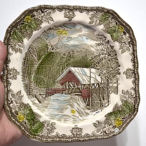 May include: A square ceramic plate with a scalloped edge featuring a transferware scene of a red covered bridge in a snowy winter landscape. The border is decorated with embossed green leaves and yellow flowers on a cream-colored background.