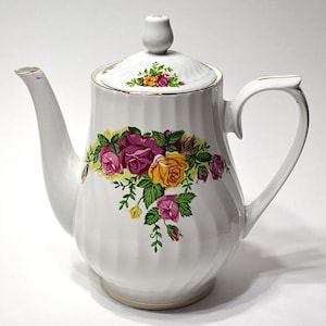 May include: A white porcelain teapot featuring a ribbed body and a floral pattern of yellow and magenta roses with green leaves. The teapot includes a matching lid with a knob handle, a curved spout, and a gold-trimmed rim and base.