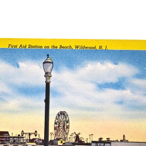 Op de afbeelding: Vintage ansichtkaart met het First Aid Station on the Beach in Wildwood, N.J. De afbeelding toont een pier met een reuzenrad, windmolens en een wapperende Amerikaanse vlag onder een blauwe lucht met wolken. Een hoge lantaarnpaal staat op de voorgrond.
