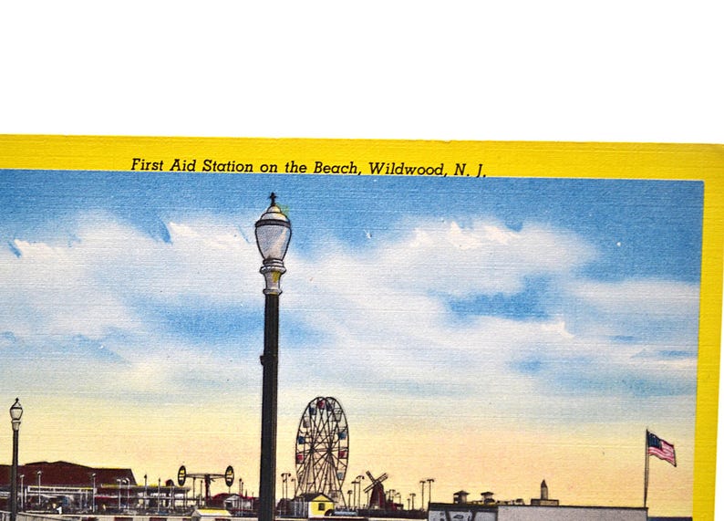 Op de afbeelding: Vintage ansichtkaart met het First Aid Station on the Beach in Wildwood, N.J. De afbeelding toont een pier met een reuzenrad, windmolens en een wapperende Amerikaanse vlag onder een blauwe lucht met wolken. Een hoge lantaarnpaal staat op de voorgrond.