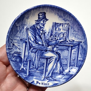May include: Blue and white ceramic plate featuring a detailed illustration of a man seated at a table, examining items. The plate has a rim with a decorative border. The word "Pedlar" is written at the bottom. The design is reminiscent of antique transferware.