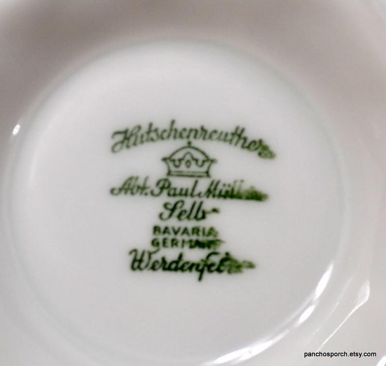 May include: Close-up of a white porcelain dish with green text and a crown logo. The text reads "Hutschenreuther Abt. Paul M&uuml;ller Selb Bavaria Germany Werdenfels."
