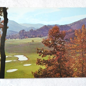 May include: A postcard depicting a picturesque view of a golf course set in a valley, surrounded by autumnal foliage. The course is green with several sand traps and a pond. The mountains in the background are covered in trees with vibrant red, orange, and yellow leaves.