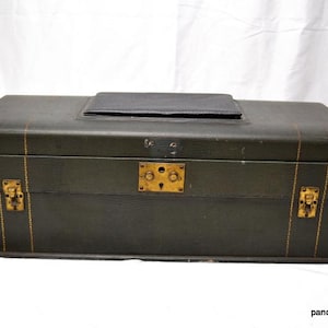 May include: A dark green vintage trunk with gold-colored hardware and stitching. The trunk has a rectangular shape with a slightly curved top and a small, black leather patch on top. The trunk is closed with two latches and a central lock.