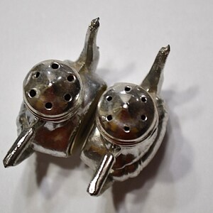 May include: A pair of silver-colored salt and pepper shakers. Each shaker is shaped like an elephant with a round top featuring multiple holes for dispensing. The shakers have a handle-like design and are set against a white background.