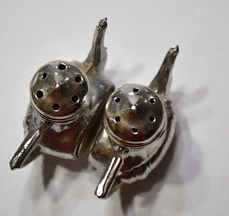 May include: A pair of silver-colored salt and pepper shakers. Each shaker is shaped like an elephant with a round top featuring multiple holes for dispensing. The shakers have a handle-like design and are set against a white background.