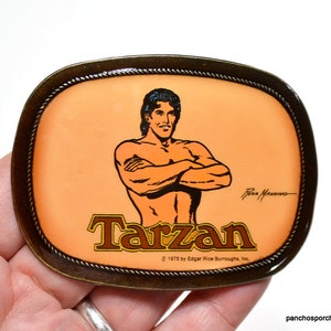 May include: An oval-shaped brass belt buckle featuring a central illustration of Tarzan with arms crossed. The background is a light orange color, and the text reads Tarzan, Ross Manning, and 1975 by Edgar Rice Burroughs, Inc. The border has a rope-like texture.
