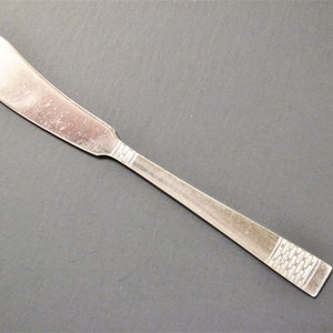 May include: A silver butter knife with a rounded blade and a rectangular handle. The handle features a decorative pattern near the end. The knife is set against a dark gray background.