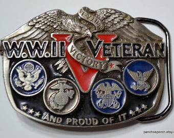 Puede incluir: Una hebilla de cinturón de metal plateado con las palabras "W.W.II VETERAN" y "VICTORY" en negro y rojo. Presenta emblemas militares en círculos azules y el texto "AND PROUD OF IT".