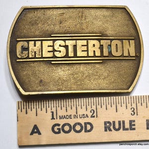 May include: A vintage brass belt buckle with the name "CHESTERTON" in raised lettering. The buckle is rectangular with rounded ends and measures approximately 3 inches in length. The words "MADE IN USA" are visible on a wooden ruler.