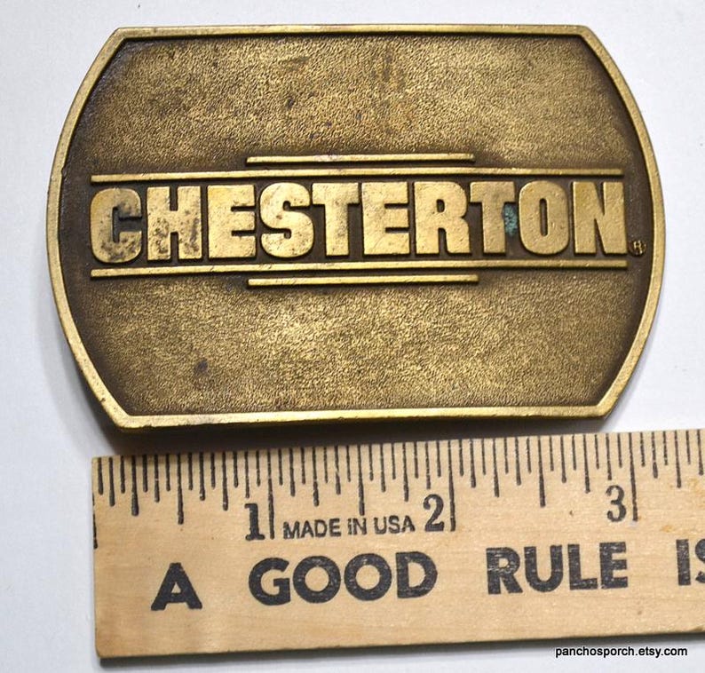 May include: A vintage brass belt buckle with the name "CHESTERTON" in raised lettering. The buckle is rectangular with rounded ends and measures approximately 3 inches in length. The words "MADE IN USA" are visible on a wooden ruler.