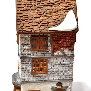 May include: A ceramic miniature house with a brown tiled roof and brick facade. Features include wooden shutters, a chimney, and a sign that reads "BATH". The house is decorated with snow accents, creating a winter scene.