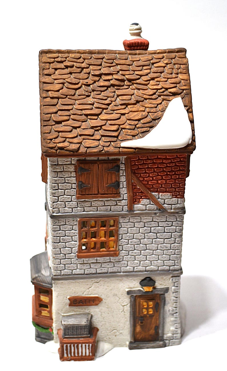 May include: A ceramic miniature house with a brown tiled roof and brick facade. Features include wooden shutters, a chimney, and a sign that reads "BATH". The house is decorated with snow accents, creating a winter scene.