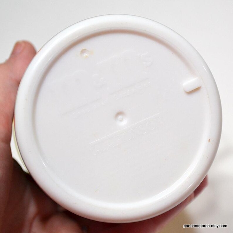 May include: A white plastic lid with a circular design. The lid has a raised edge and a small tab. The bottom of the lid has embossed text and a small hole. The lid is held by a hand.