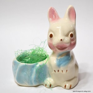 May include: A vintage ceramic bunny planter with a light blue planter base. The bunny has pink accents on its ears and cheeks, with brown eyes and a blue bow. The planter is filled with green decorative grass.