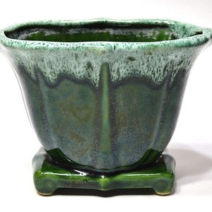May include: A green ceramic planter with a unique, fluted design. The planter has a square base and a flared top with a textured, two-tone glaze in shades of green and white. The planter is suitable for indoor or outdoor use.